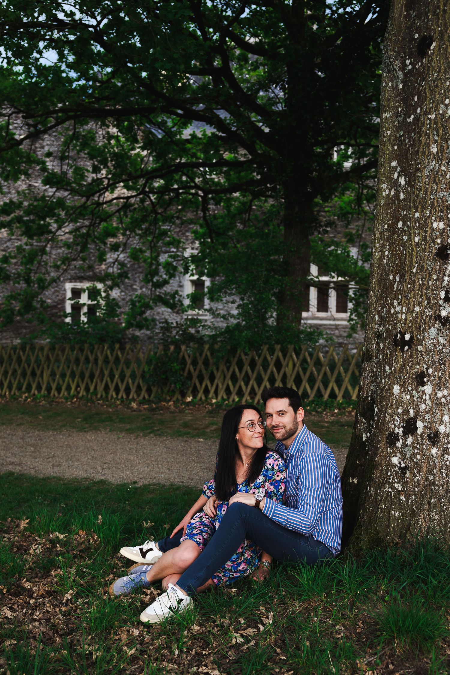 Couple sitting together against a tree trunk in a lush green park, woman in floral dress, man in blue striped shirt.