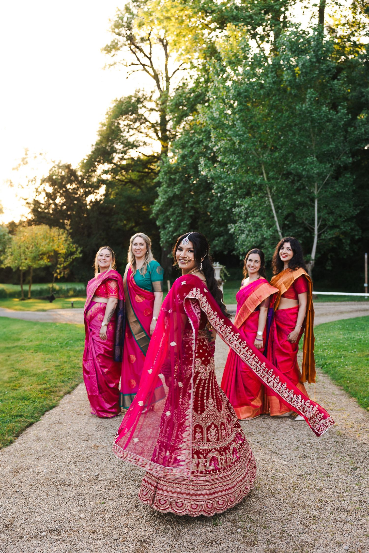 Five women in vibrant red and pink traditional South Asian lehengas sit together on a grassy path at sunset.
