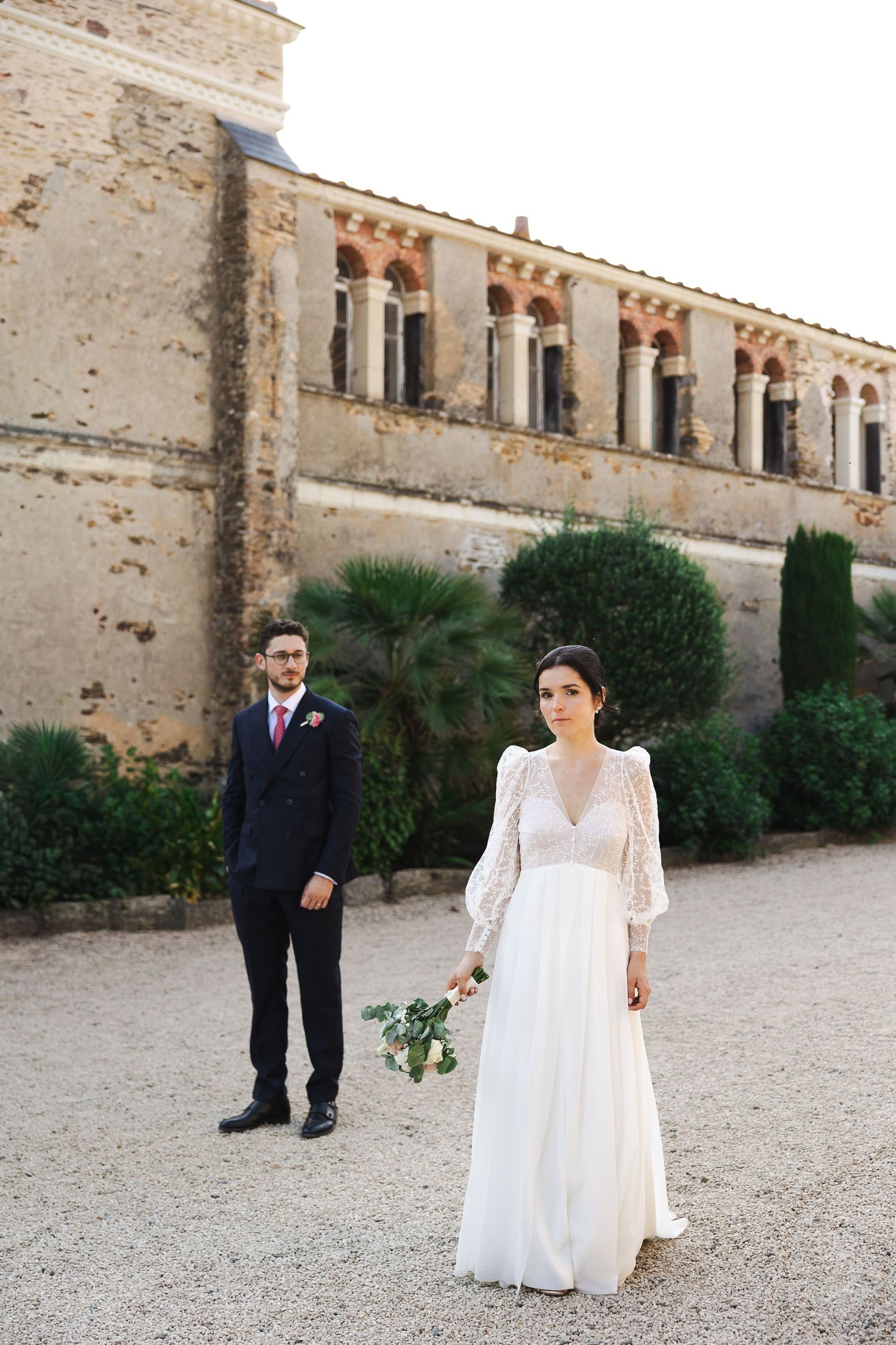 Bride and groom posing outside historic stone building, bride holding bouquet in lace gown.