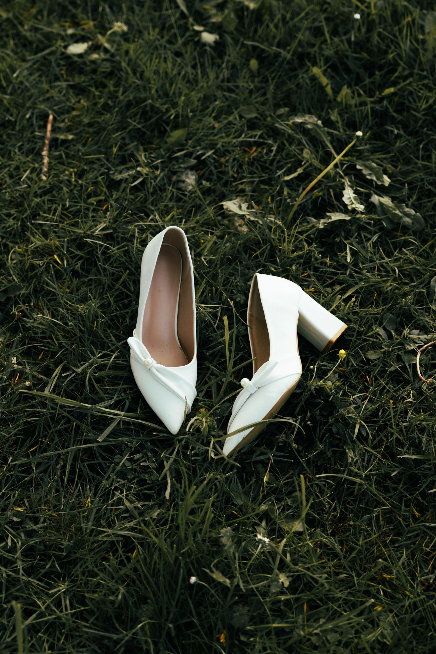 Elegant white bridal heels with pointed toes lying on dark green grass.