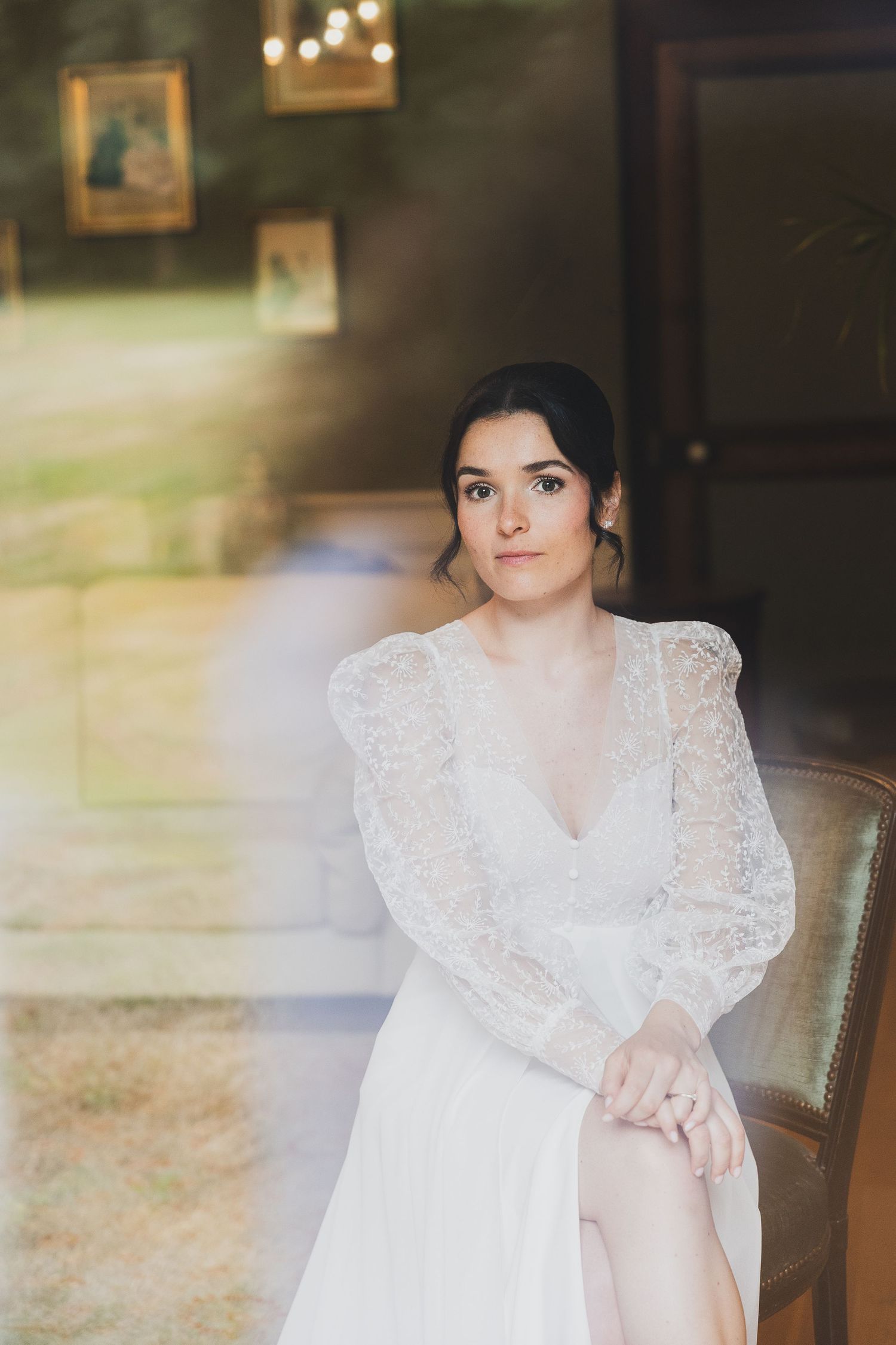 Elegant bride in lace long-sleeve white wedding dress seated in vintage chair indoors.
