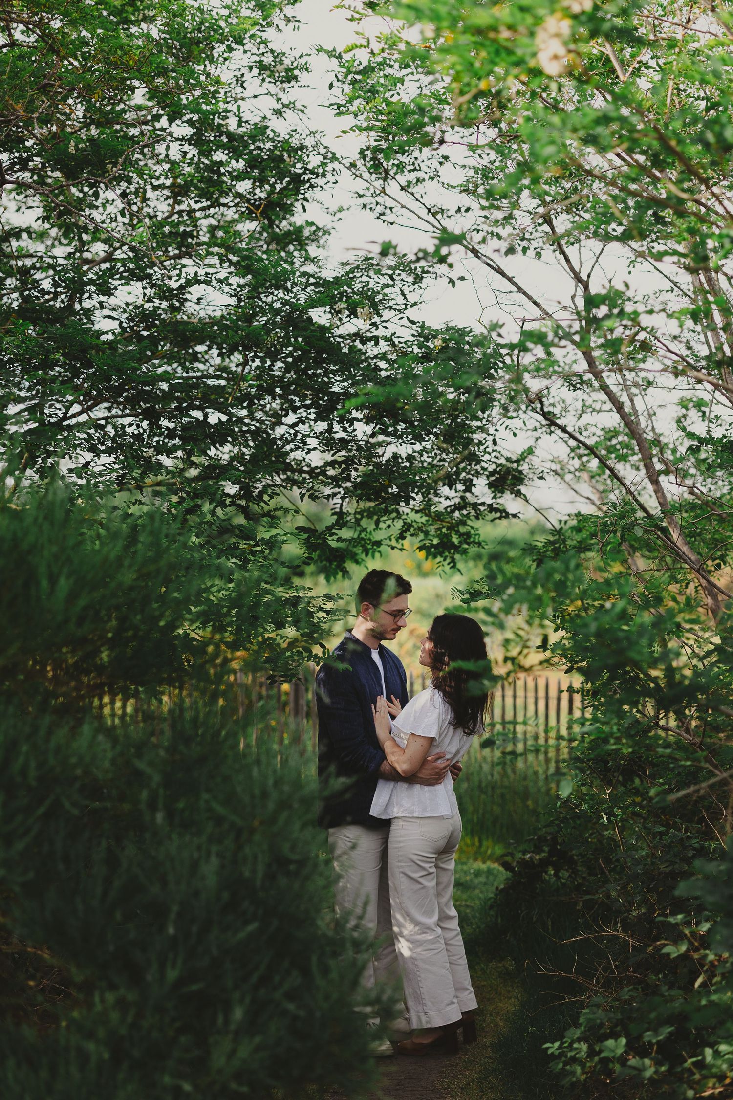 A couple embraces romantically among lush green garden trees and shrubs near a wooden fence.