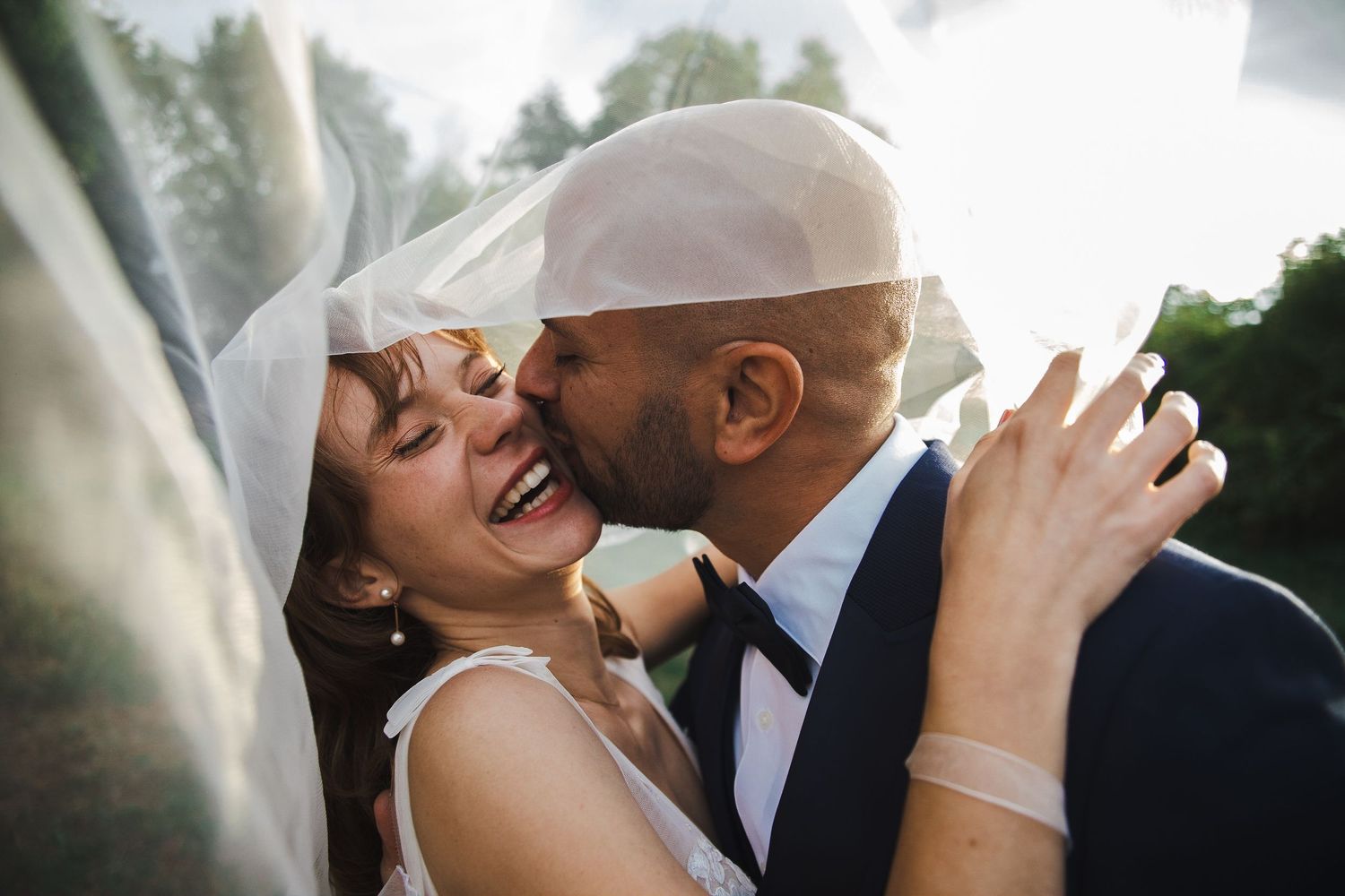 A couple shares an intimate wedding kiss outdoors while being photographed from multiple romantic angles.
