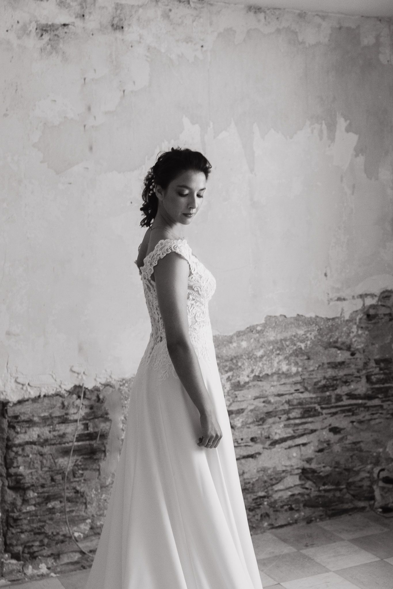 A bride in a romantic lace wedding gown poses elegantly against a distressed wall in black and white photography.