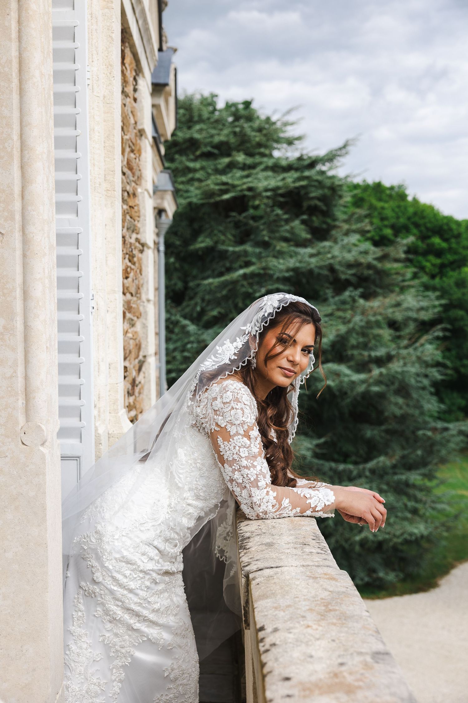 A bride in a long-sleeved lace wedding gown and veil leans against a stone balcony with green trees in the background.