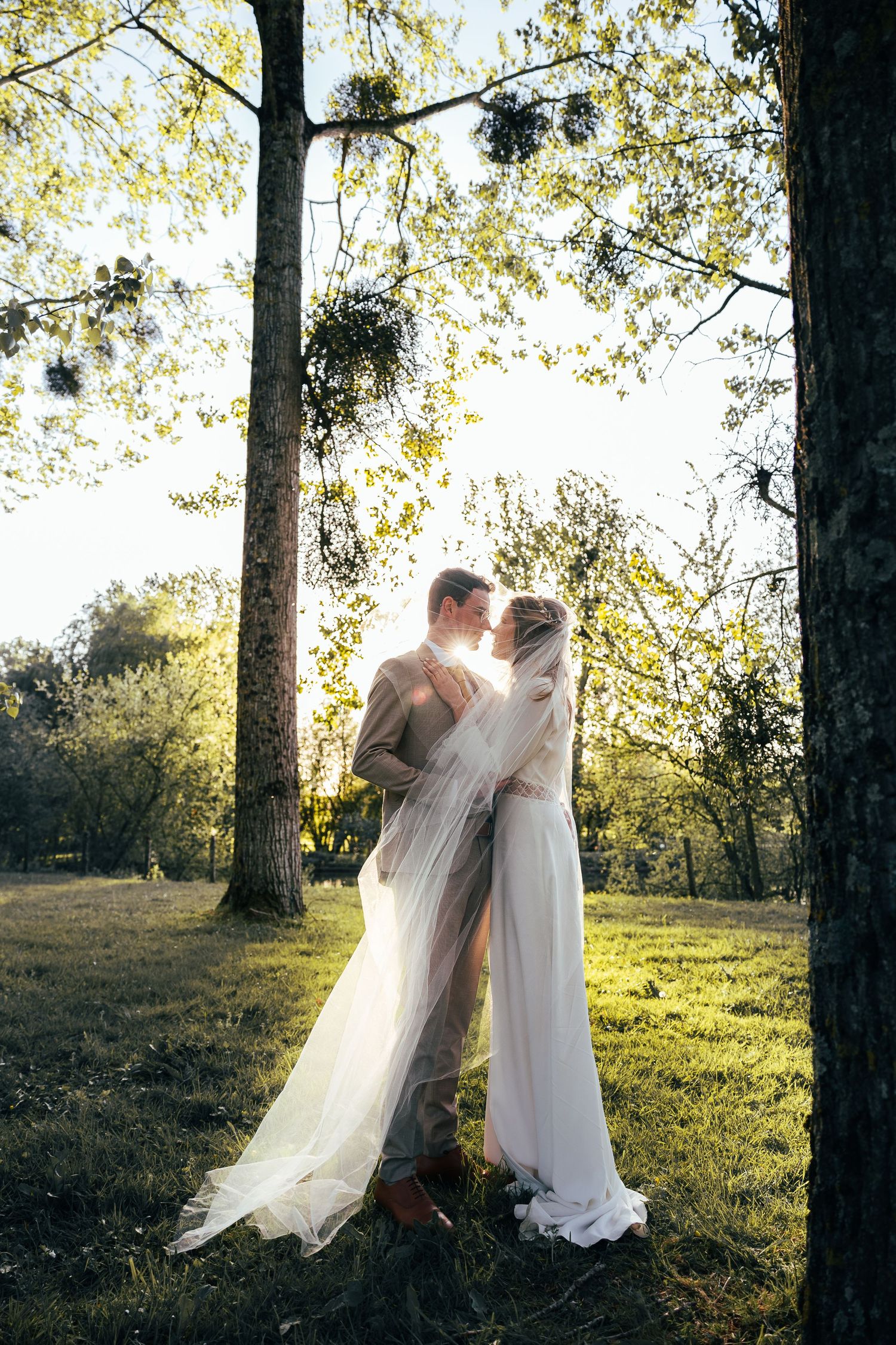A couple in flowing white attire share an intimate moment in a sunlit forest setting with tall trees.