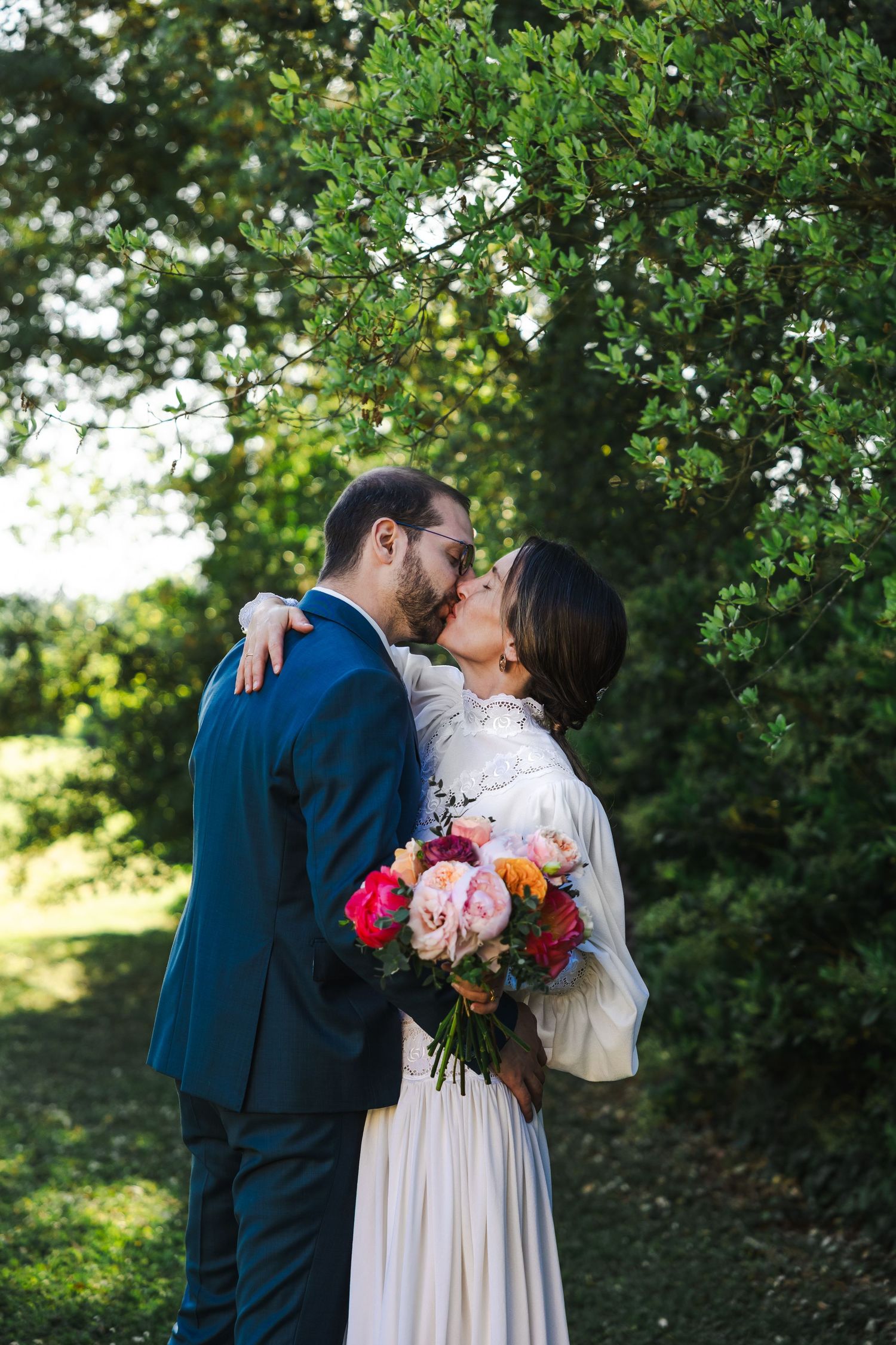Two people embrace romantically under a green tree while holding a vibrant bouquet of pink and orange flowers.