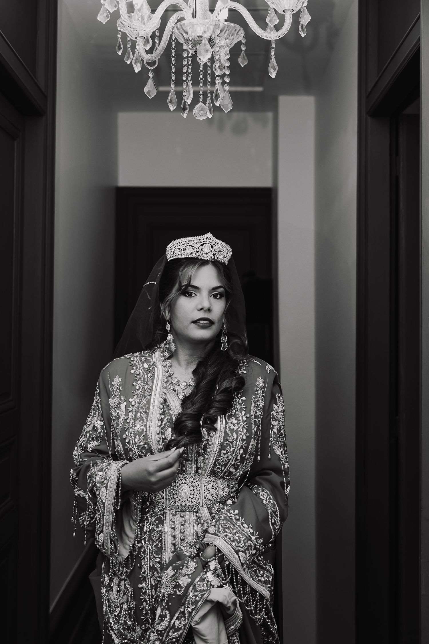 Black and white glamour portrait series showing a model in an ornate crown and embellished outfit under chandeliers.
