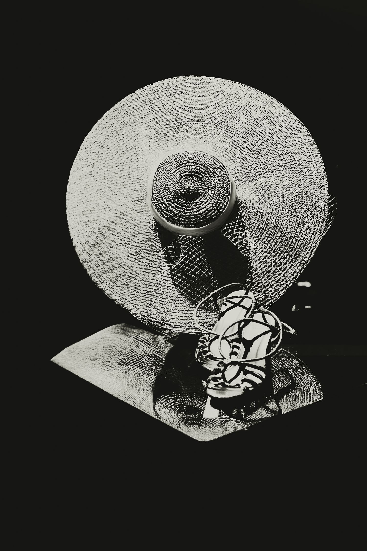 A black and white still life photograph of a wide-brimmed straw hat and decorative circular object creating dramatic shadows.