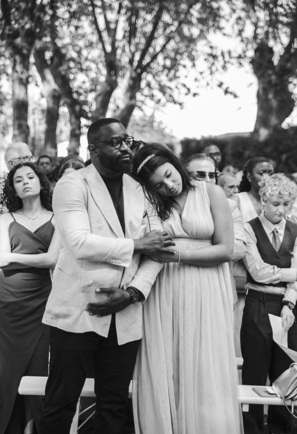 A couple in formal attire share an intimate moment at an outdoor gathering among other guests.