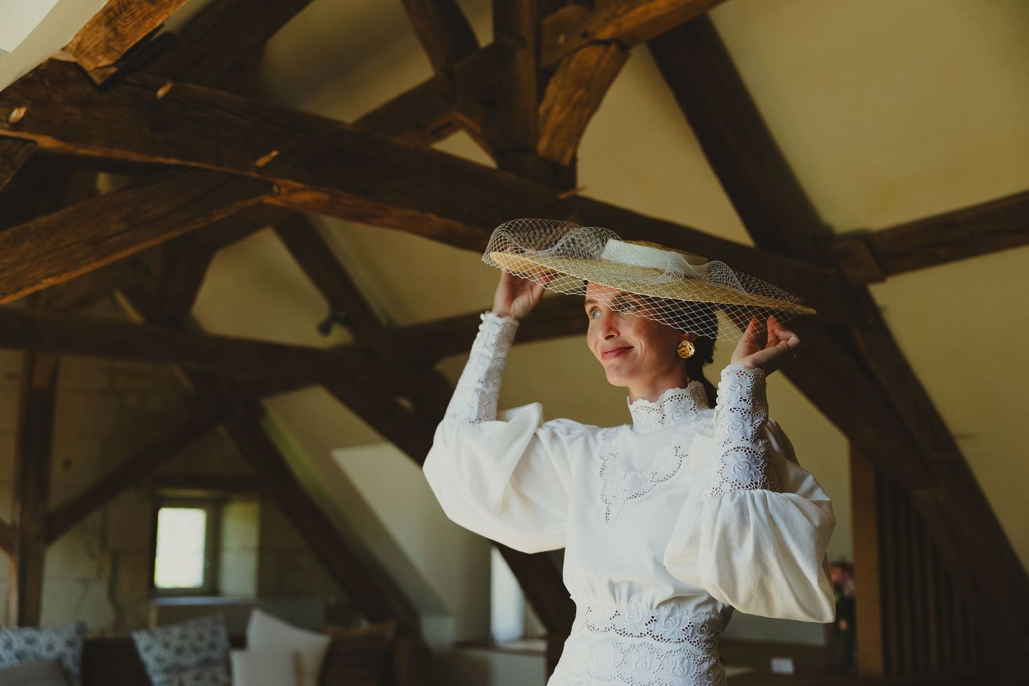 Bride in white lace dress adjusting vintage veil hat in rustic barn with exposed wooden beams.