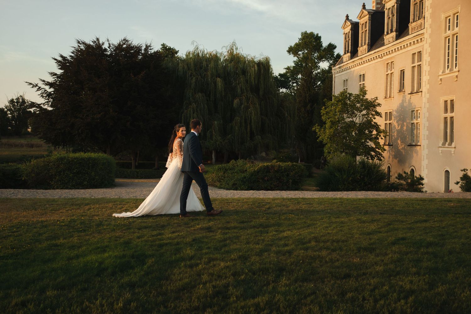A couple in wedding attire embraces on a sunlit lawn beside an elegant French chateau during golden hour.