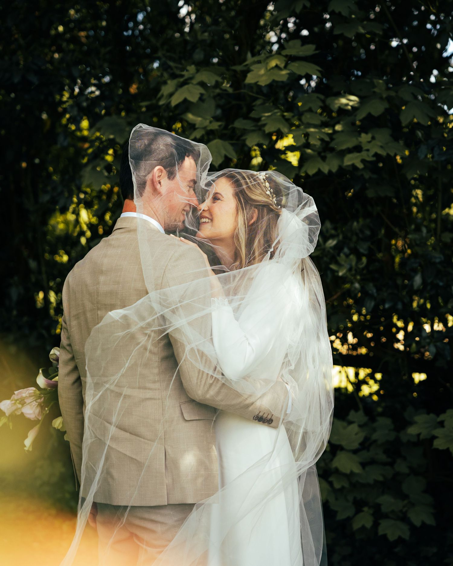 A romantic wedding couple embraces outdoors with sunlight filtering through green foliage in a garden setting.