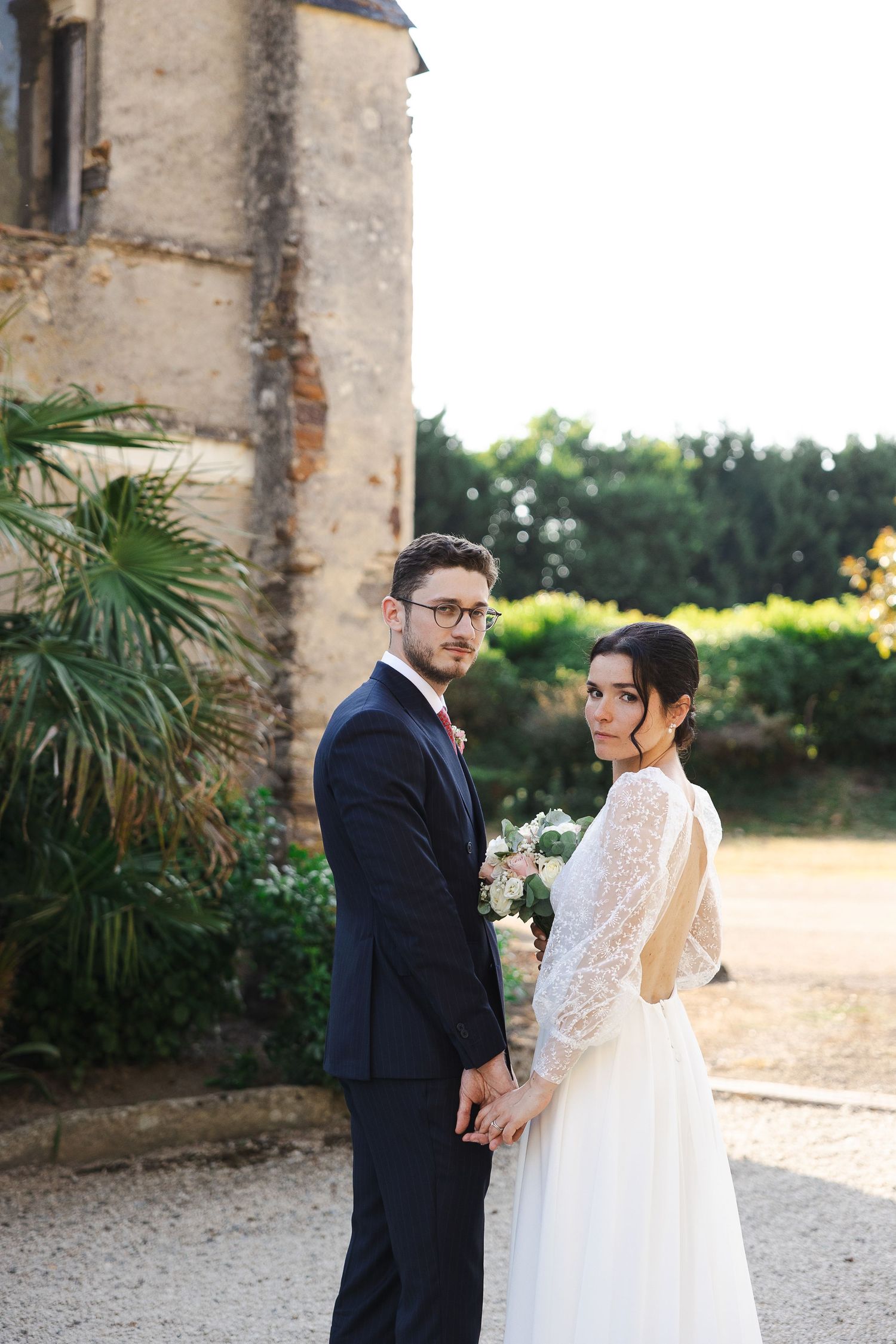 Bride and groom holding hands outdoors near a historic stone building, bride in lace dress holding bouquet.