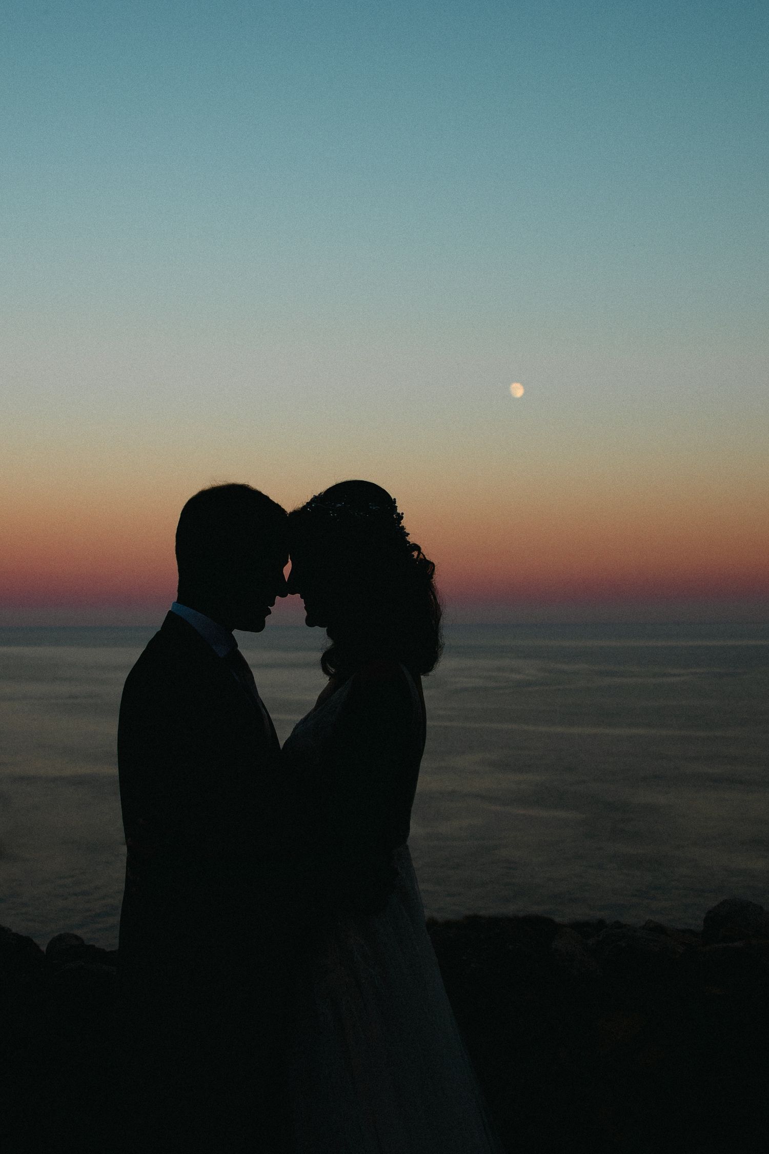 Silhouetted couple sharing an intimate moment against a sunset sky with the moon visible over the ocean.