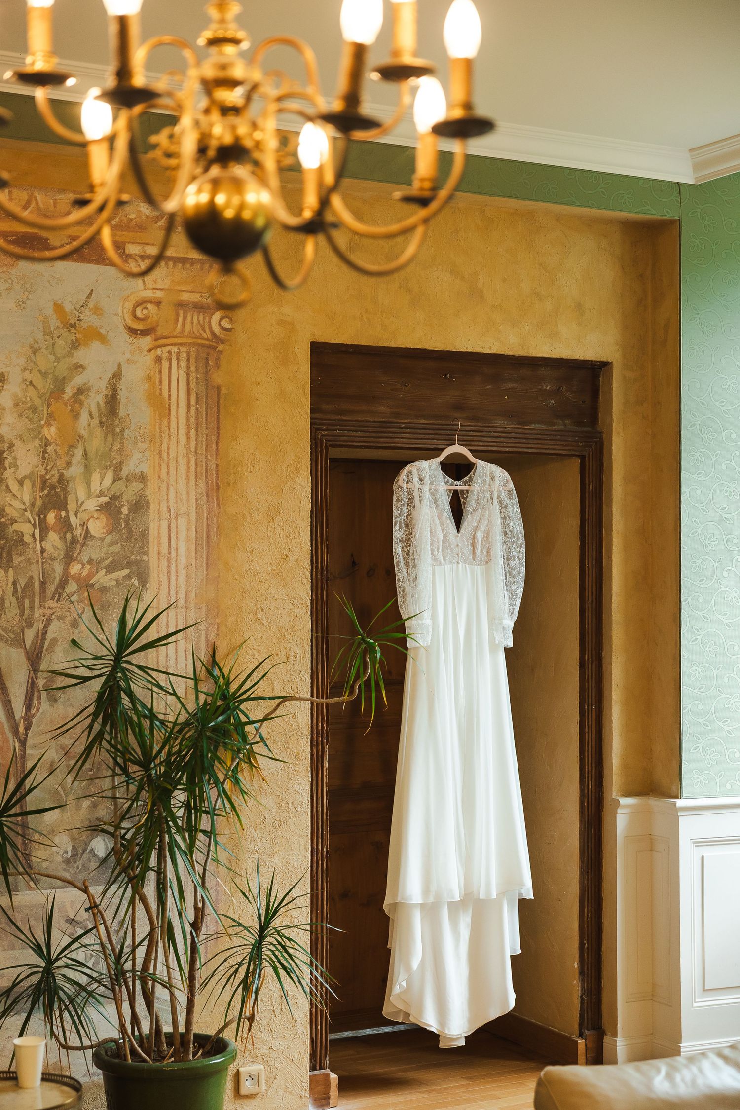 Elegant white lace wedding dress hanging on a wooden door in a vintage room with chandelier and tropical plants.