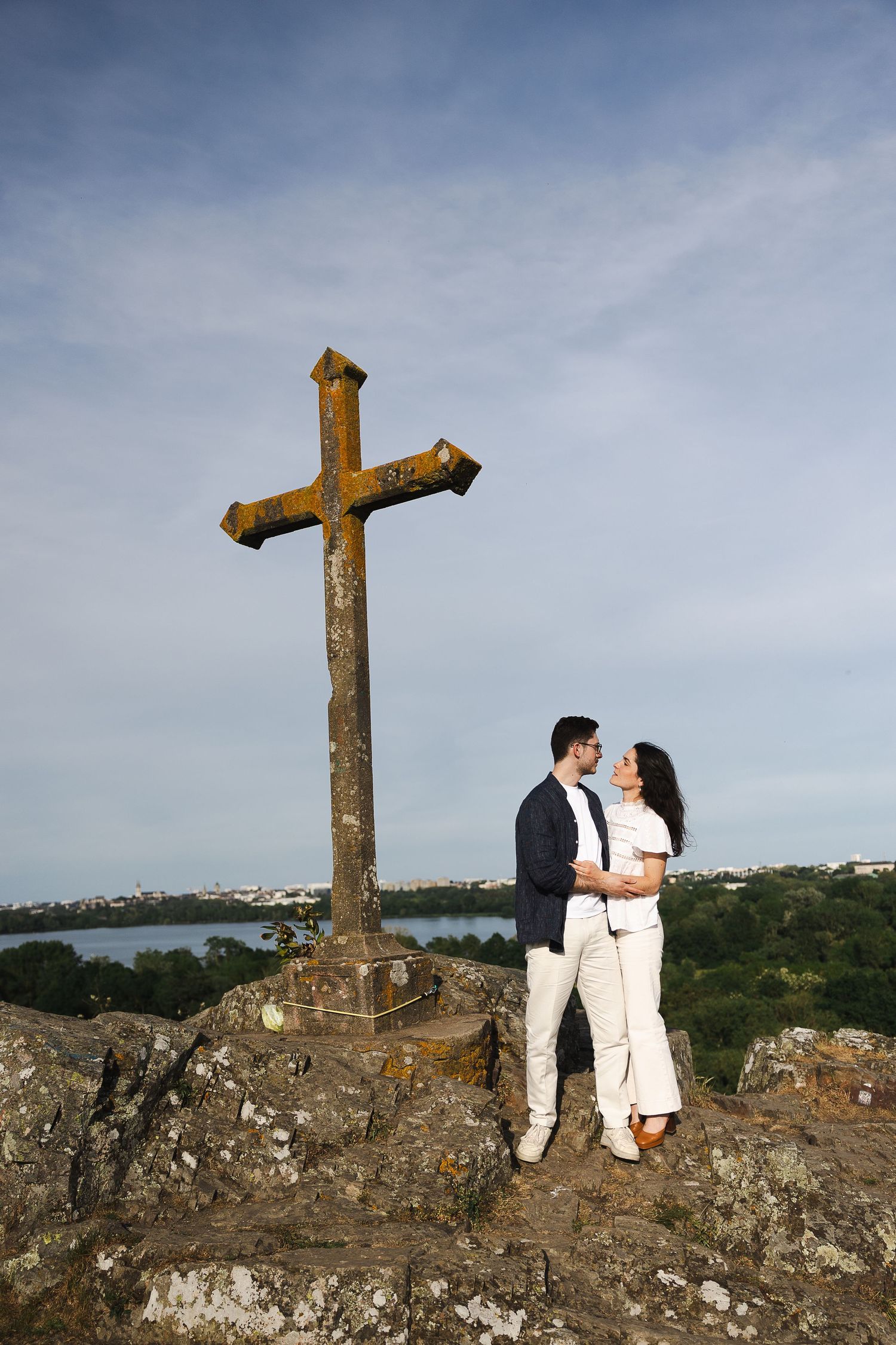 Couple embracing on rocky hilltop beside an old stone cross with scenic coastal views and blue sky.
