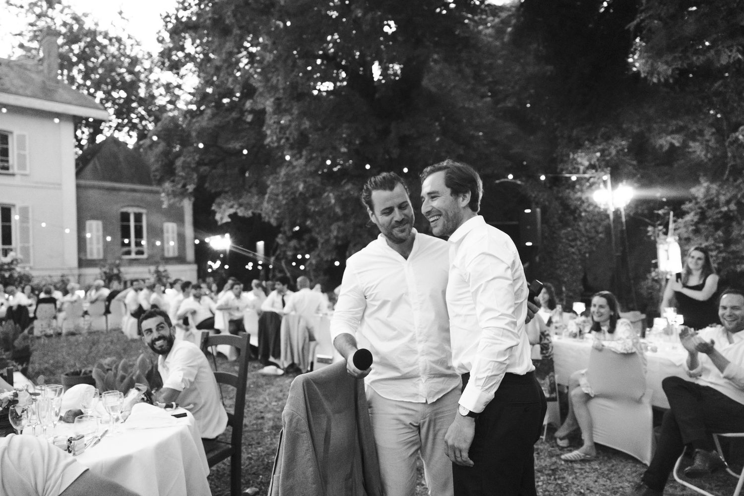 Two men in white shirts share a laugh at an elegant outdoor garden party event in black and white photo.