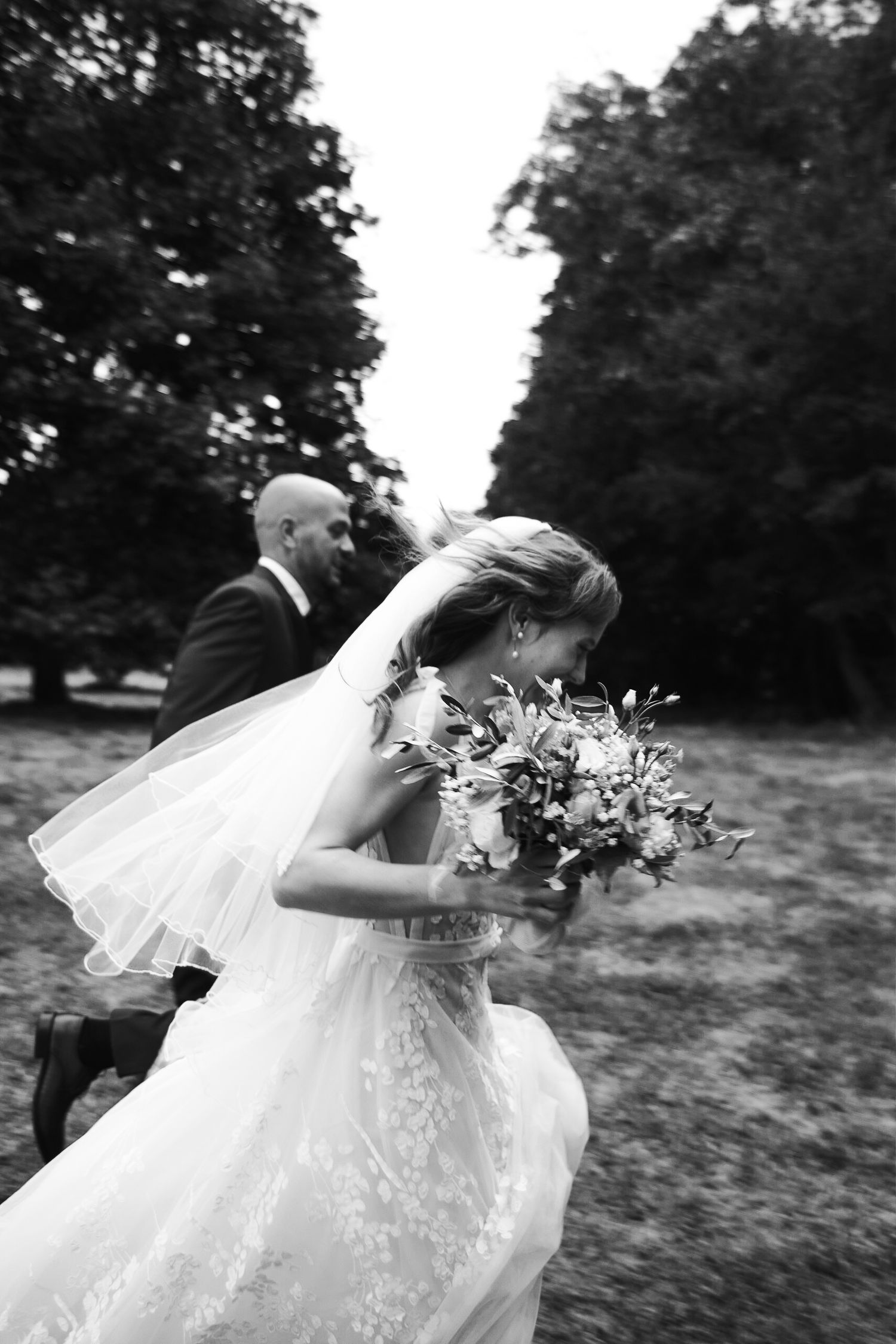 A black and white wedding photograph capturing a joyful moment as a bride in a flowing veil runs with her bouquet outdoors.