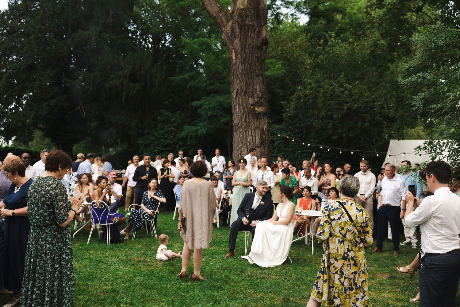 Outdoor wedding ceremony with guests gathered around bride and groom seated at a table beneath large trees.
