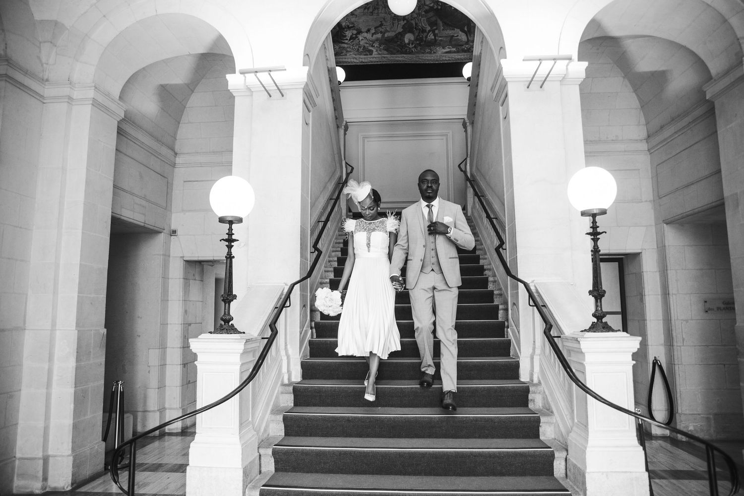 A couple in wedding attire descends a grand white staircase with elegant columns and railings in black and white.