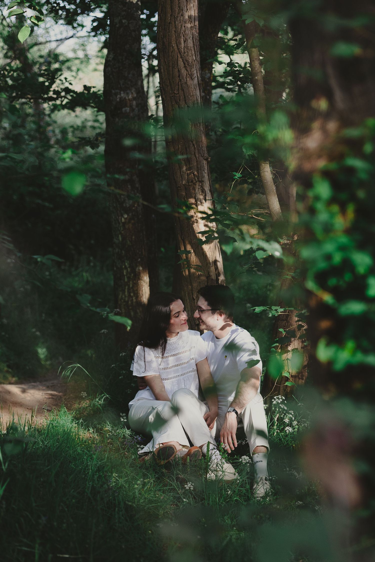 Couple sitting together in a lush green forest, wearing white outfits, sharing a romantic moment among tall trees.