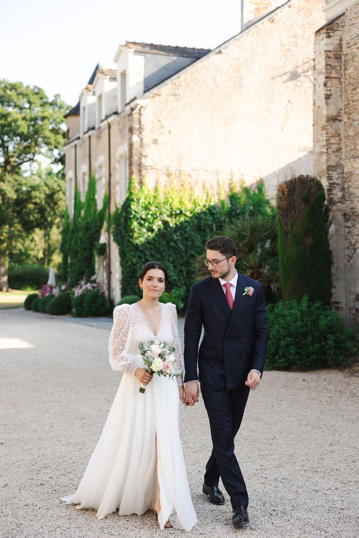 A bride and groom walk hand in hand on a gravel path beside a stone building surrounded by lush greenery.