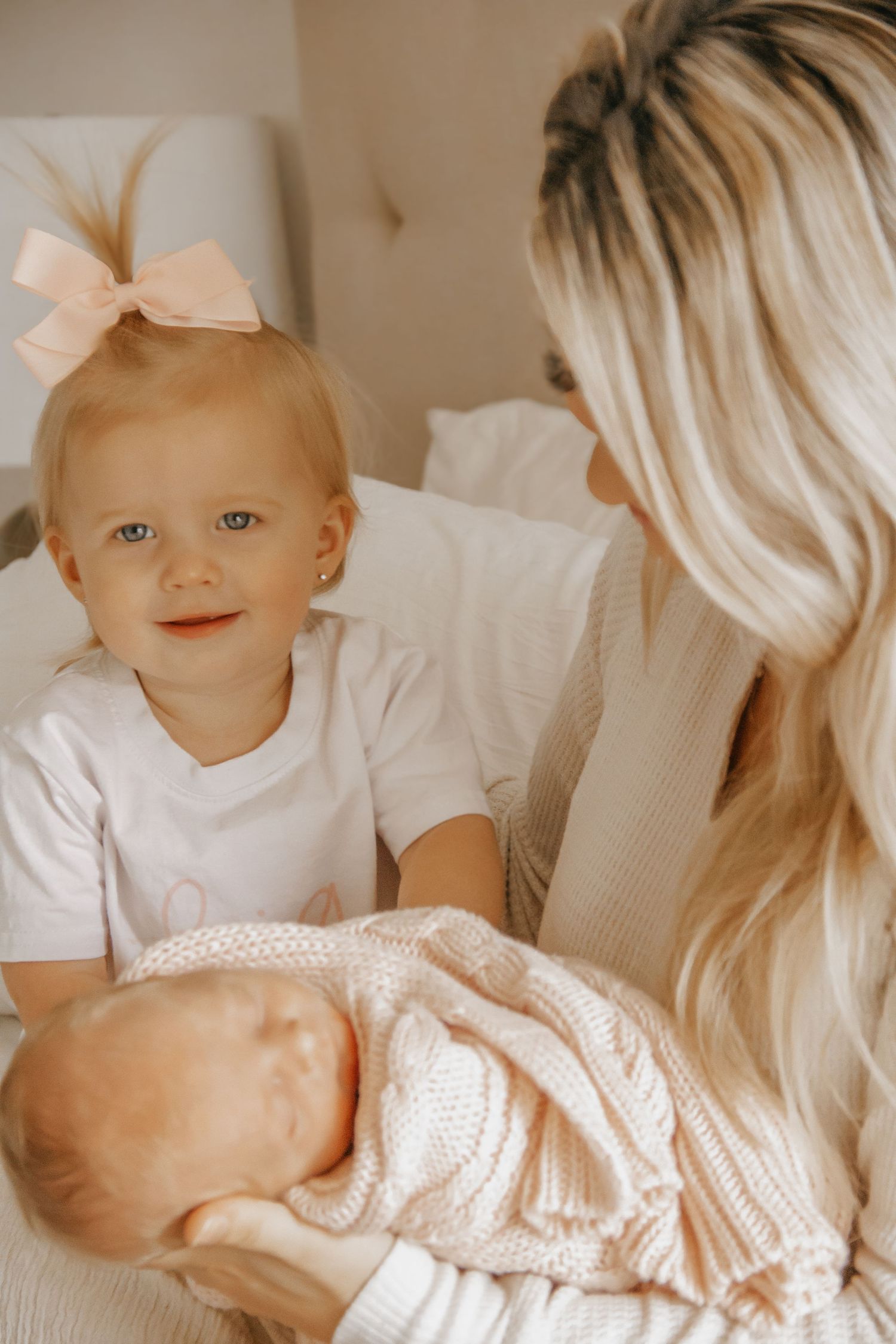Blonde mother holding newborn baby while toddler girl with pink bow looks on lovingly.