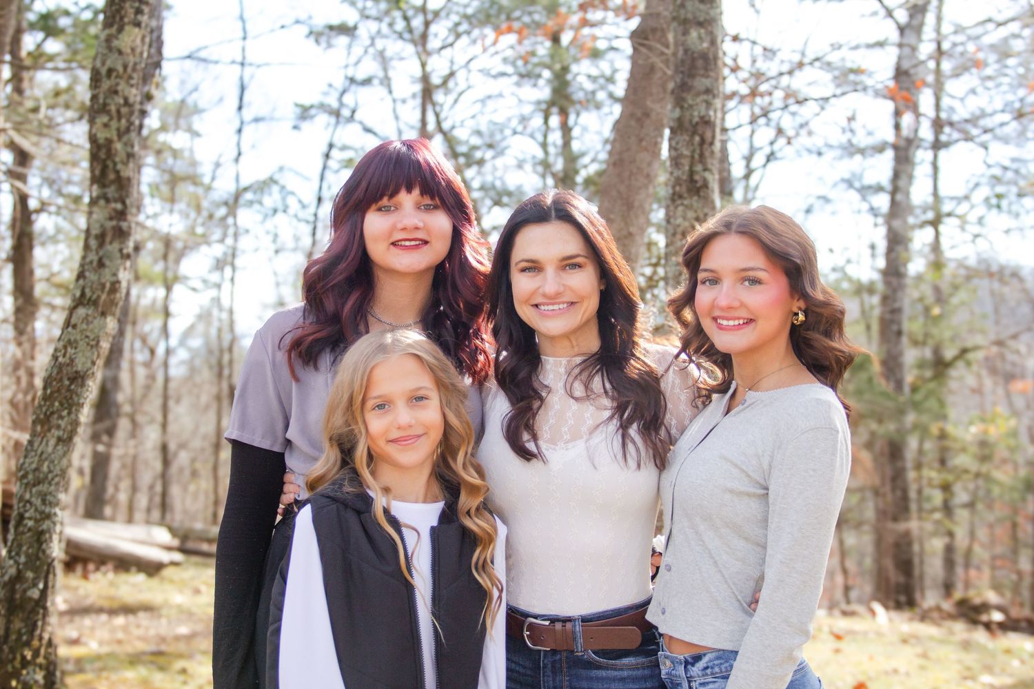 Four girls smiling together outdoors in a wooded setting, posing for a family portrait in casual attire.