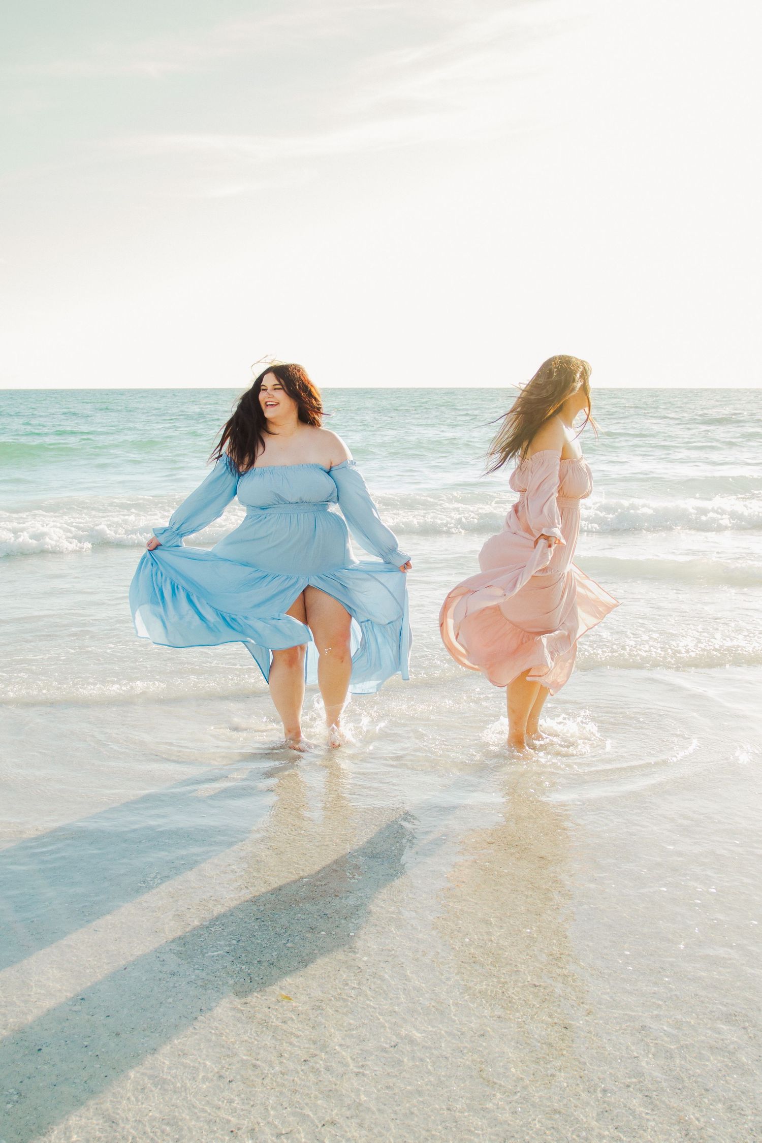 Two women in flowing blue and pink dresses walk along a sunny beach with turquoise waves.