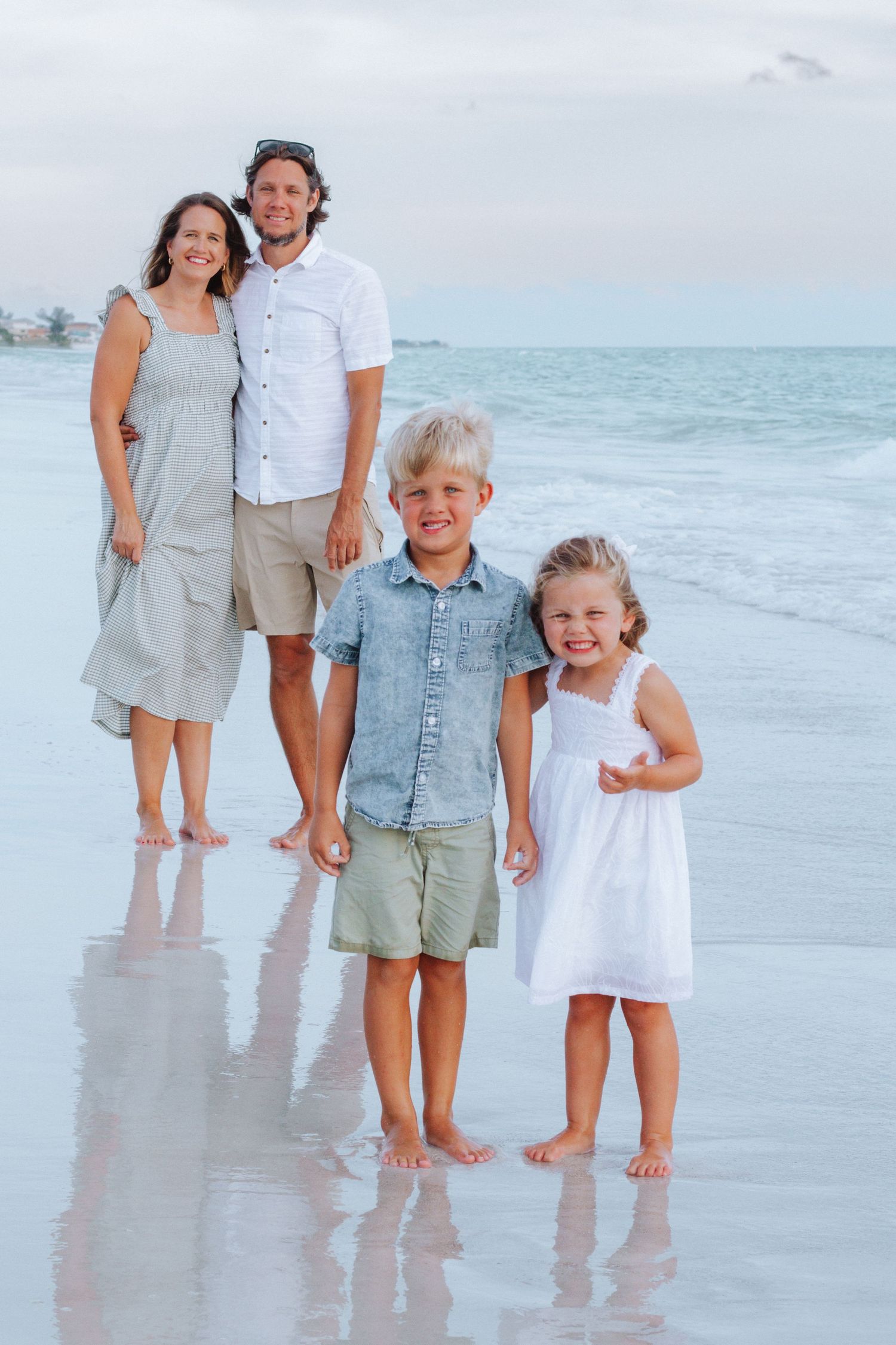 Happy family of four posing barefoot on sandy beach with ocean waves behind them.