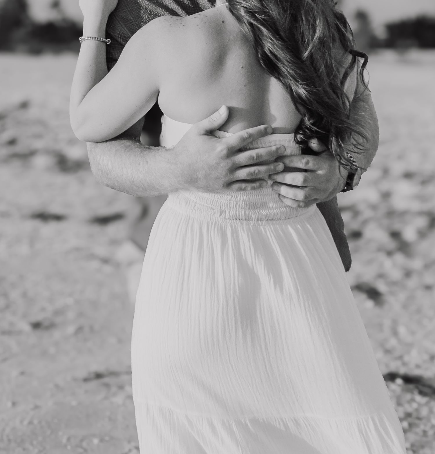 Black and white photo of a couple embracing on a beach, woman in white skirt with long hair.