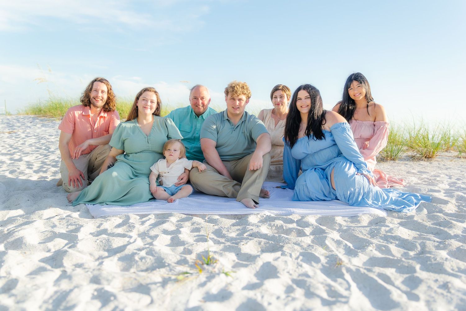 Large family group smiling together on white sand beach, wearing coordinated pastel outfits for a professional portrait.