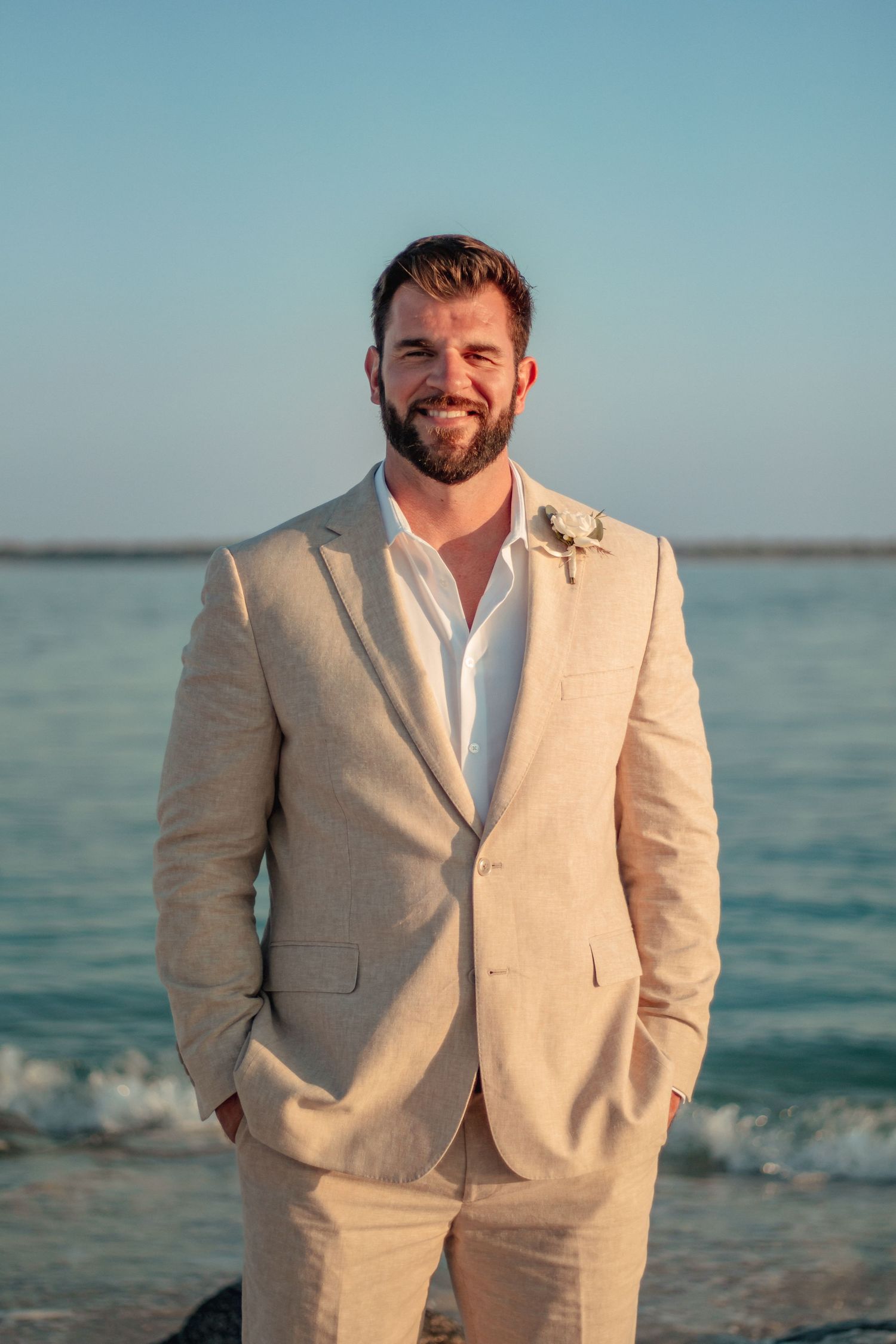 Smiling bearded groom in tan linen suit with boutonniere posing on beach at sunset.