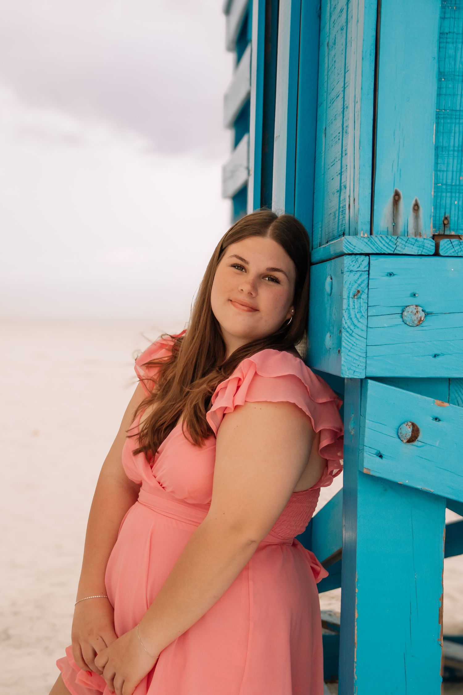 Young woman in pink ruffle dress leaning against turquoise beach hut on sandy shore.