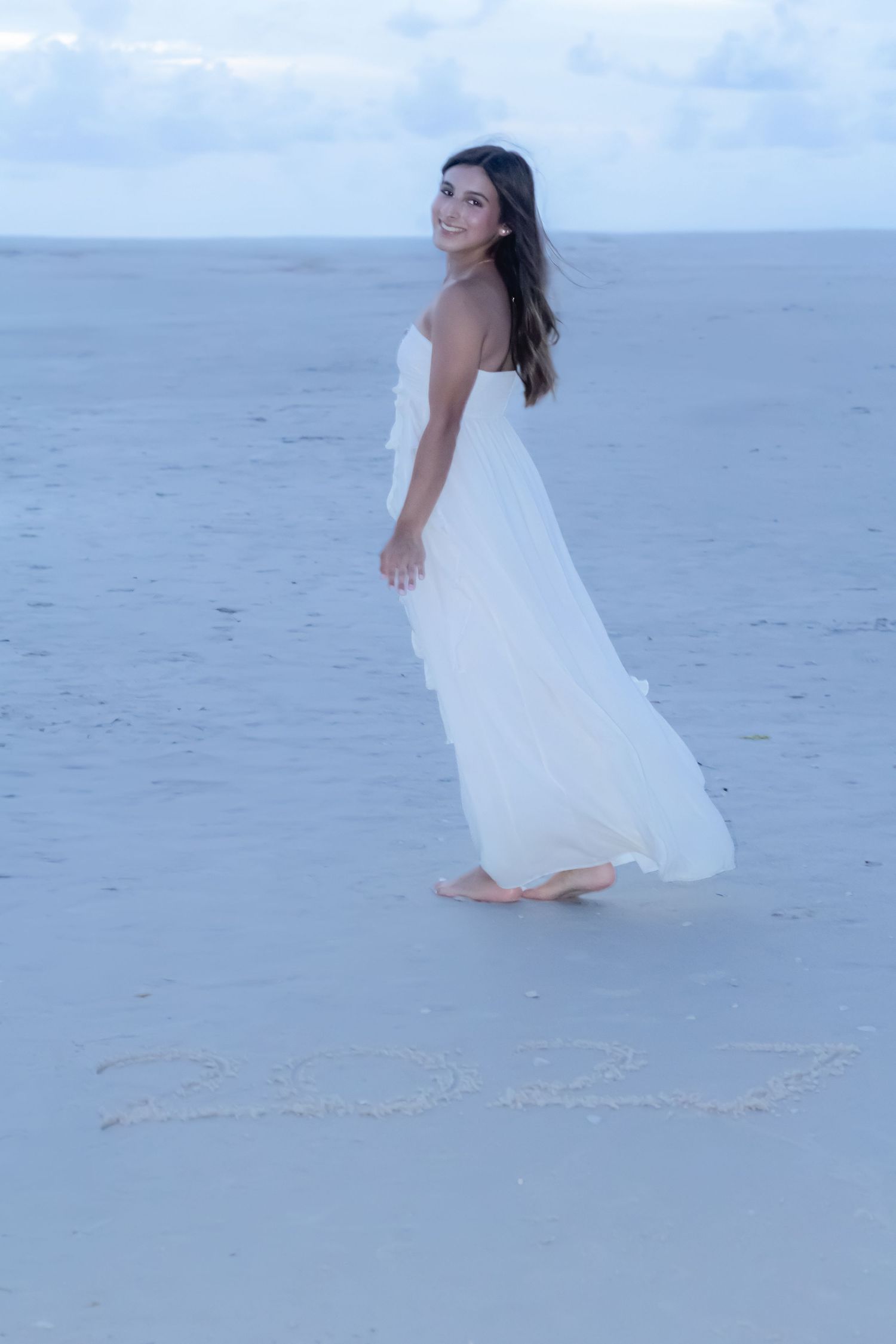 A smiling woman in a flowing white strapless dress walks barefoot on a sandy beach with a cloudy sky backdrop.