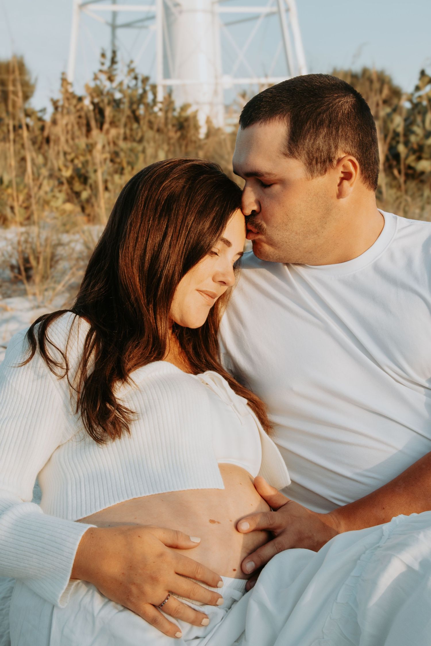 A man kisses a pregnant woman's forehead as they sit together outdoors, both wearing white, cradling her baby bump.