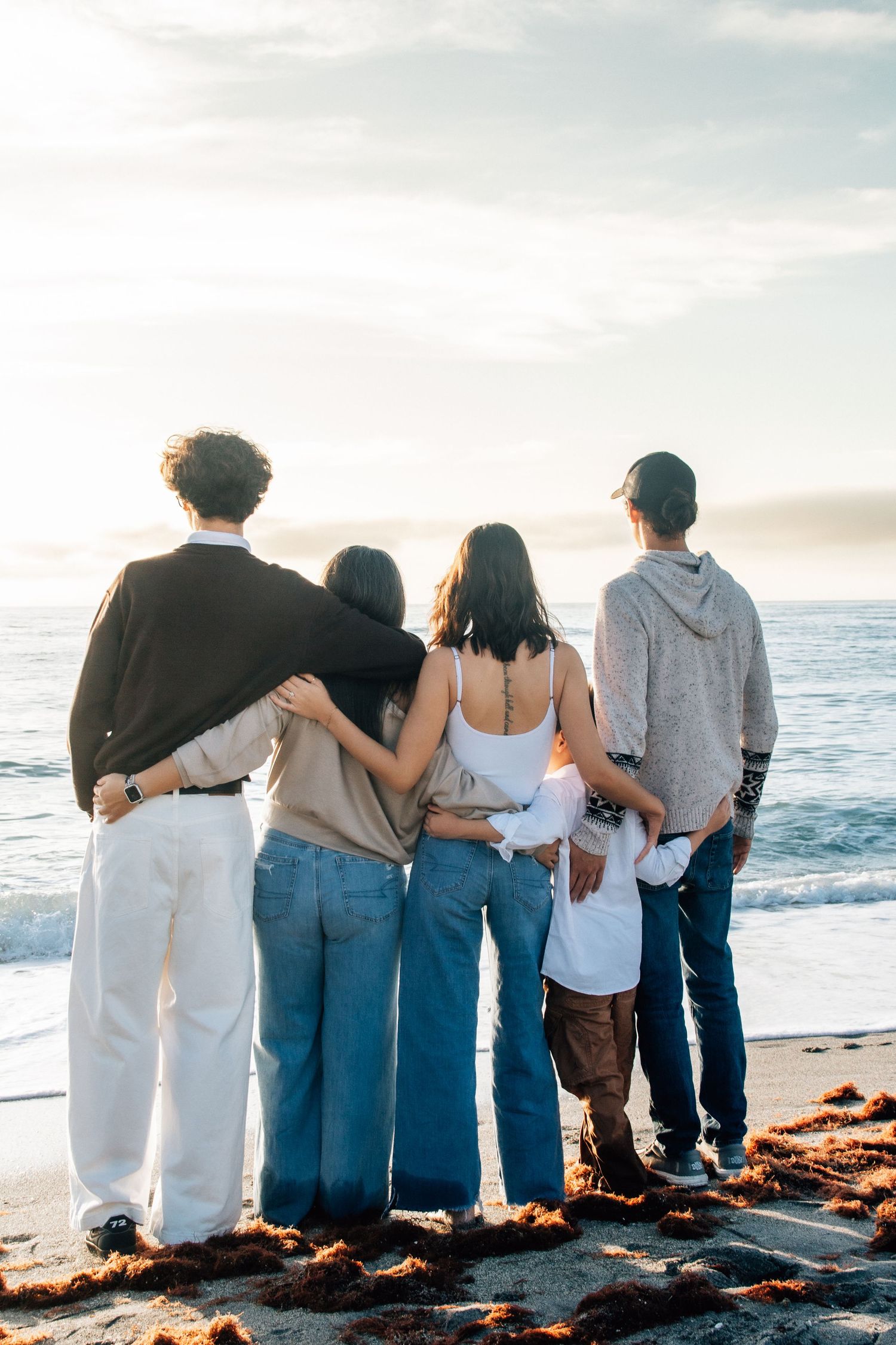 Group of five friends embracing on a beach at sunset, facing the ocean in casual jeans and tops.