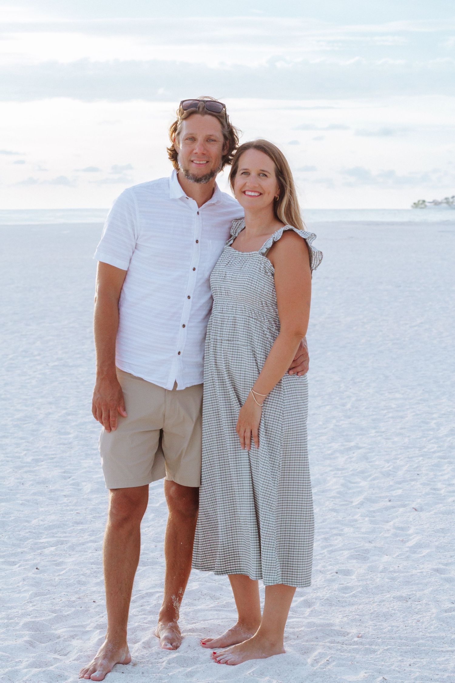 Couple smiling together on a white sand beach, man in white shirt and shorts, woman in sage green midi dress.