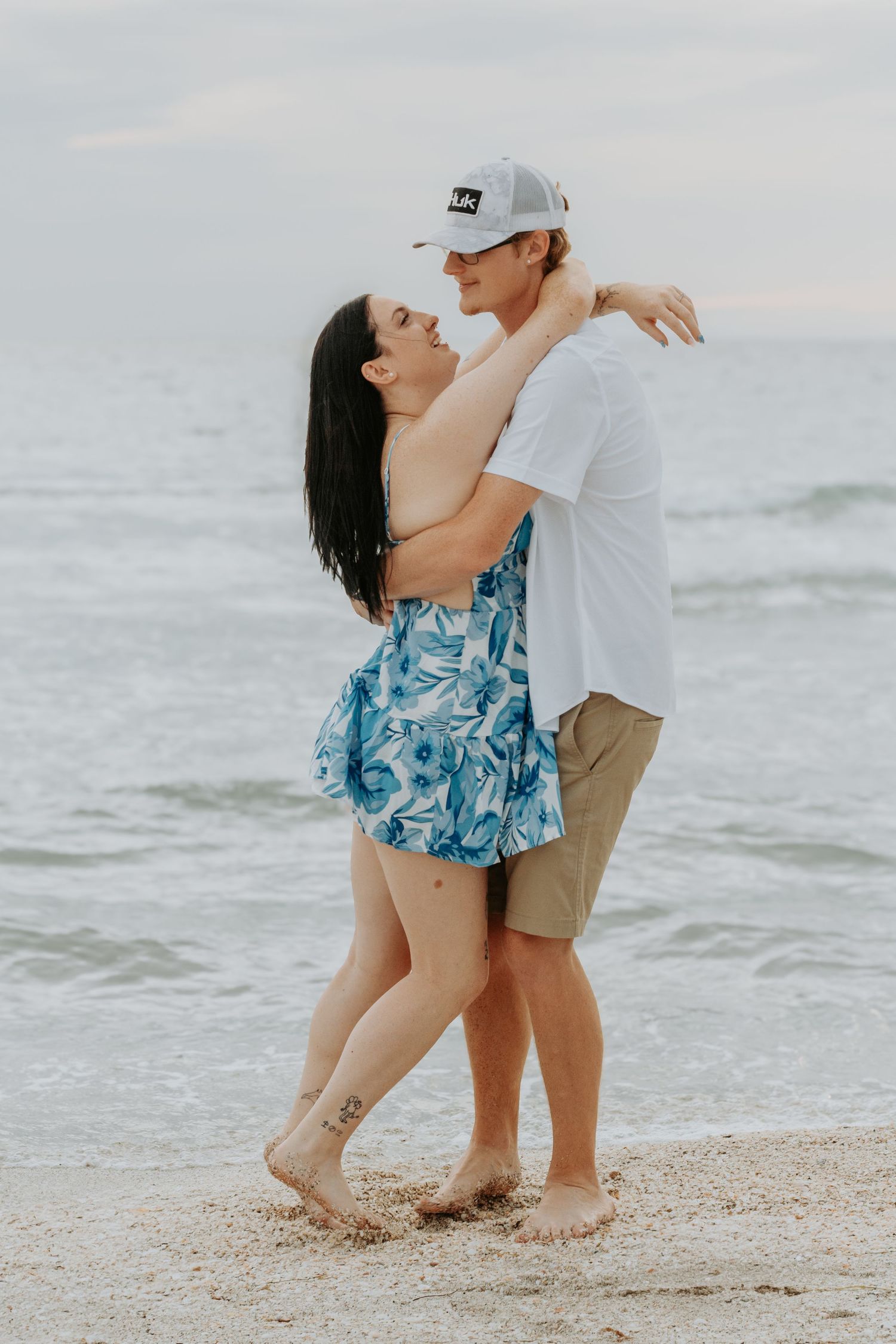Couple embracing romantically on a sandy beach, woman in blue floral dress and man in white shirt and cap.