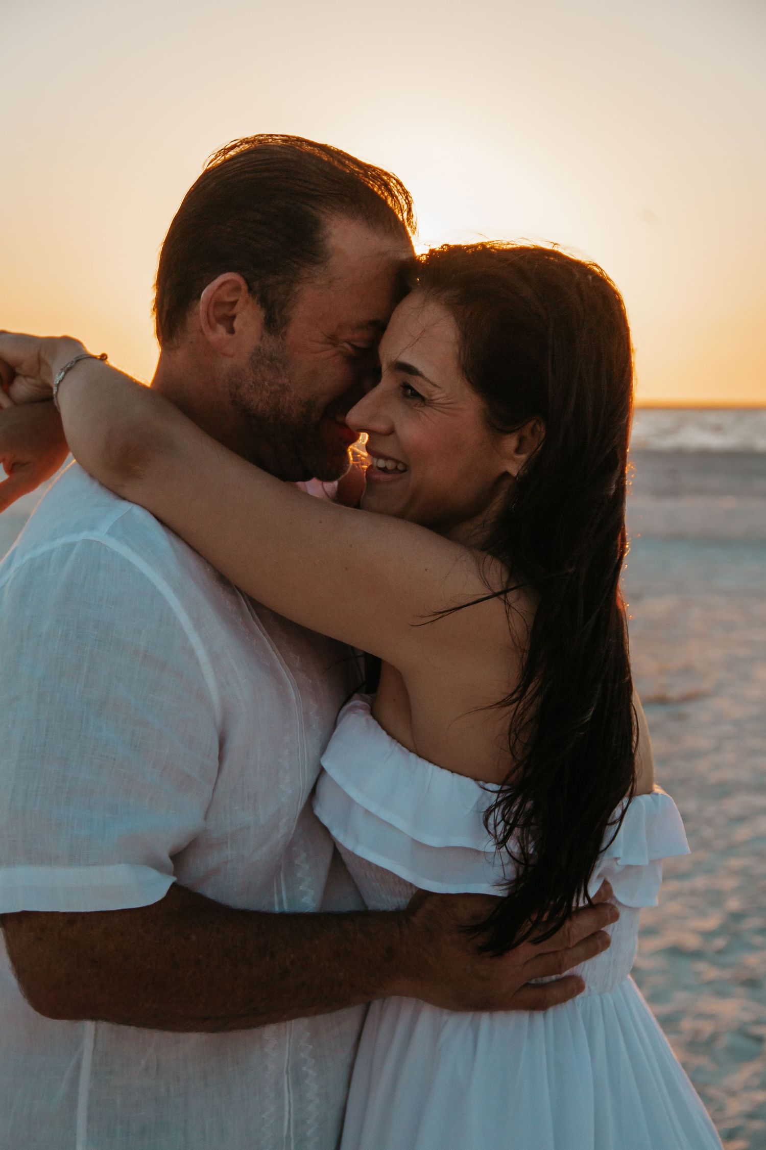Couple embracing at sunset on the beach, wearing white outfits, sharing a romantic moment by the ocean.