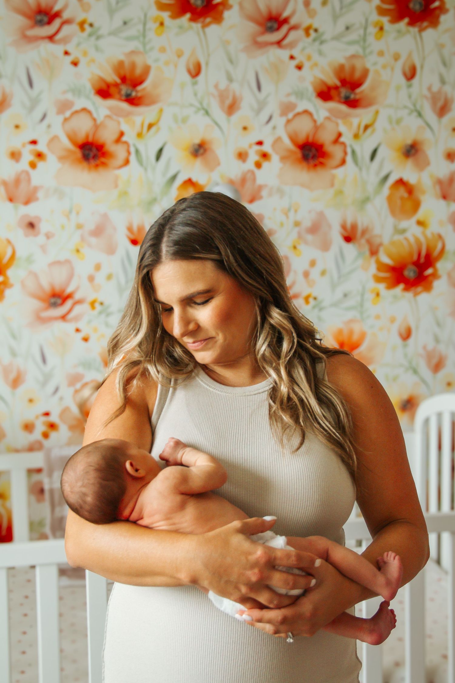A mother in a white dress tenderly holds her newborn baby in a floral nursery room.