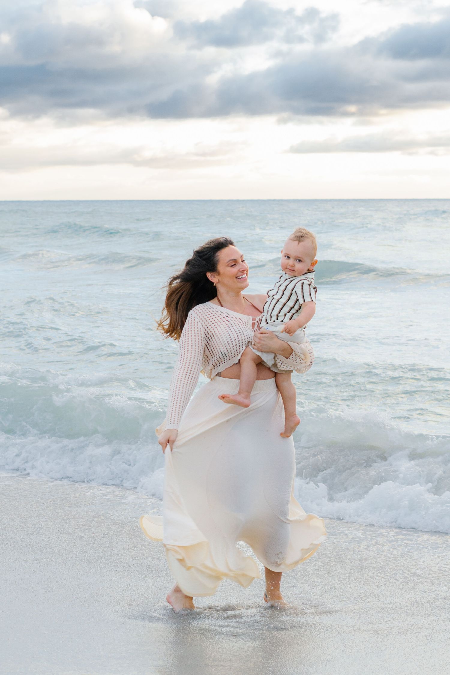A joyful mother in a white dress holds her baby on a breezy beach with ocean waves in the background.