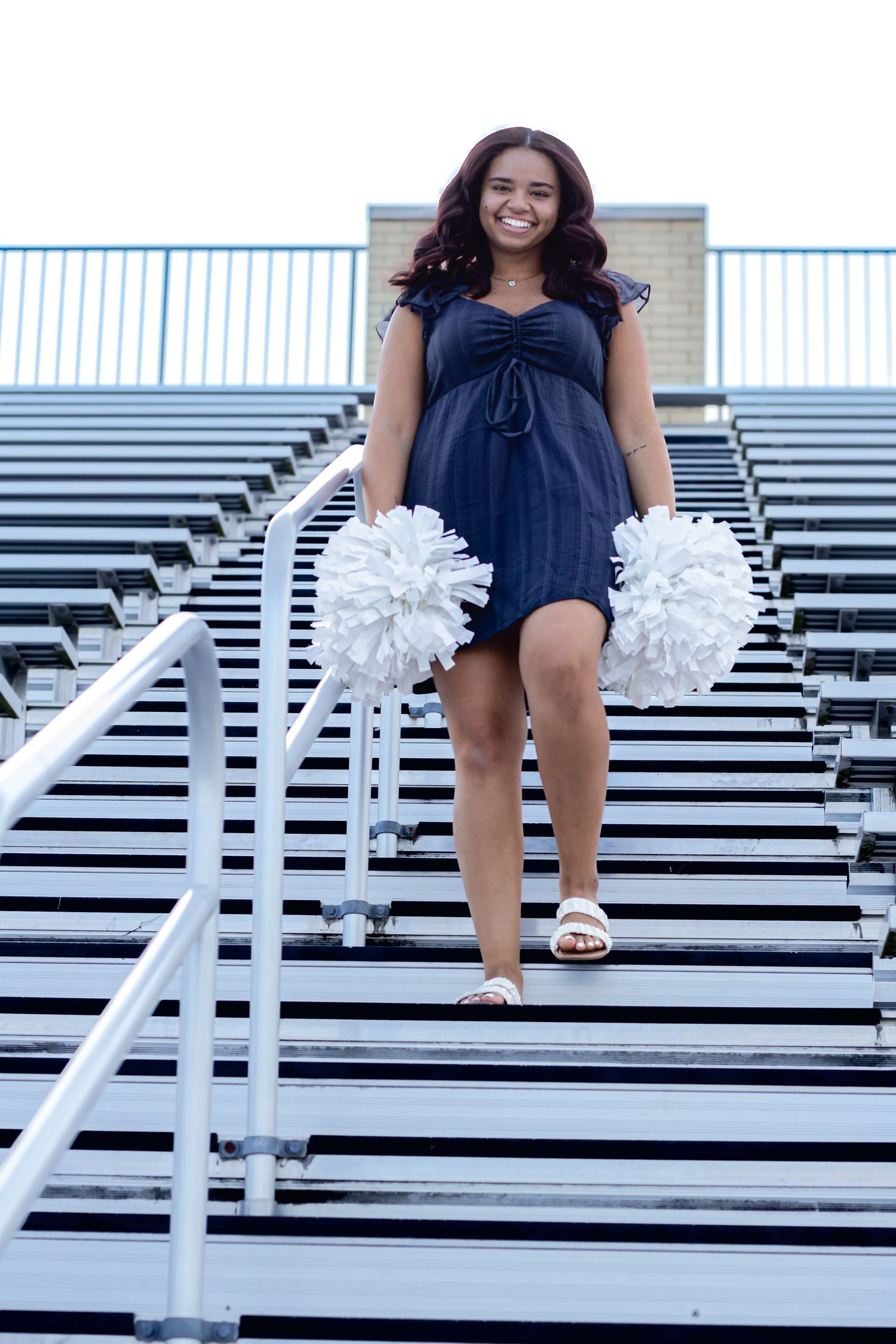 Young cheerleader in navy dress holds white pom-poms while walking down stadium bleacher steps, smiling confidently.