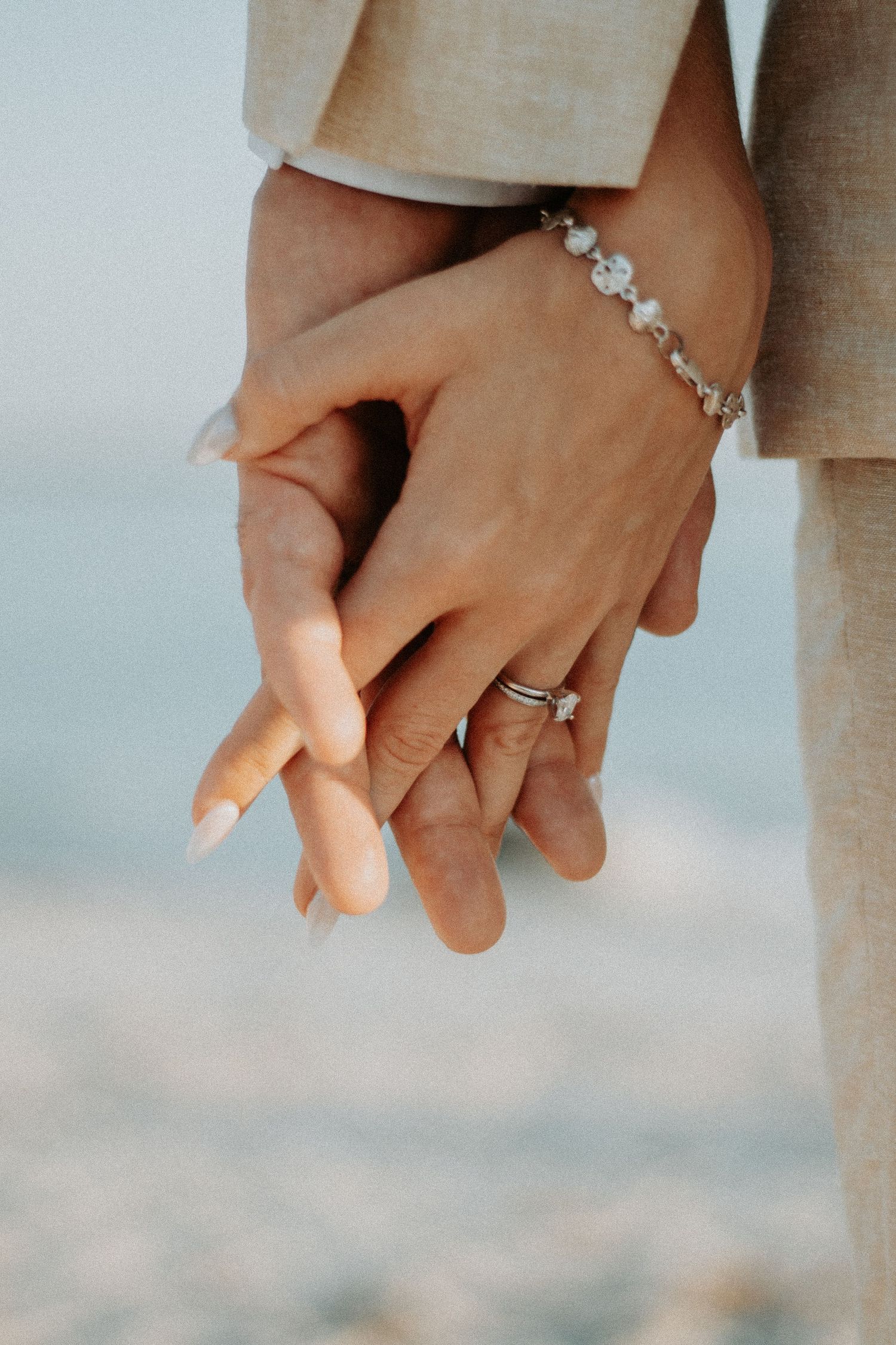 Close-up of couple holding hands on beach, woman wearing diamond engagement ring and elegant bracelet.