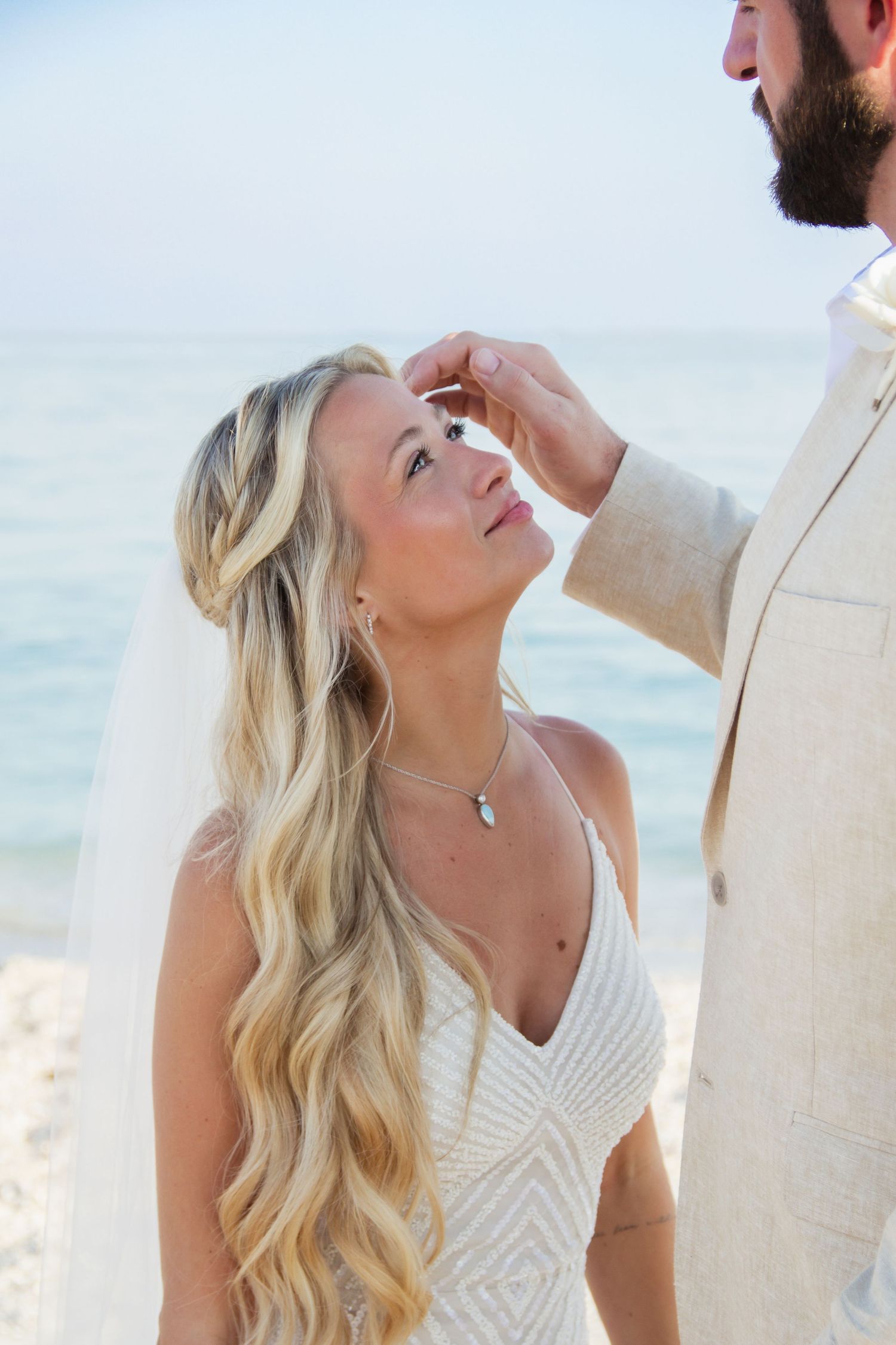 Bride in white beaded gown with flowing blonde hair gazes up at groom in tan suit during romantic beach wedding ceremony.