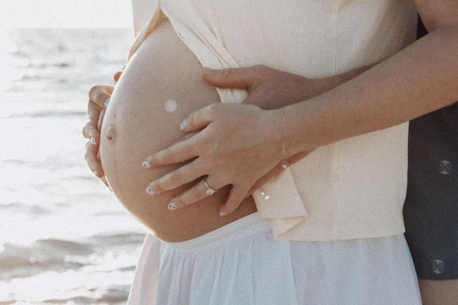 Pregnant woman in white dress with partner embracing her baby bump on a sunny beach.