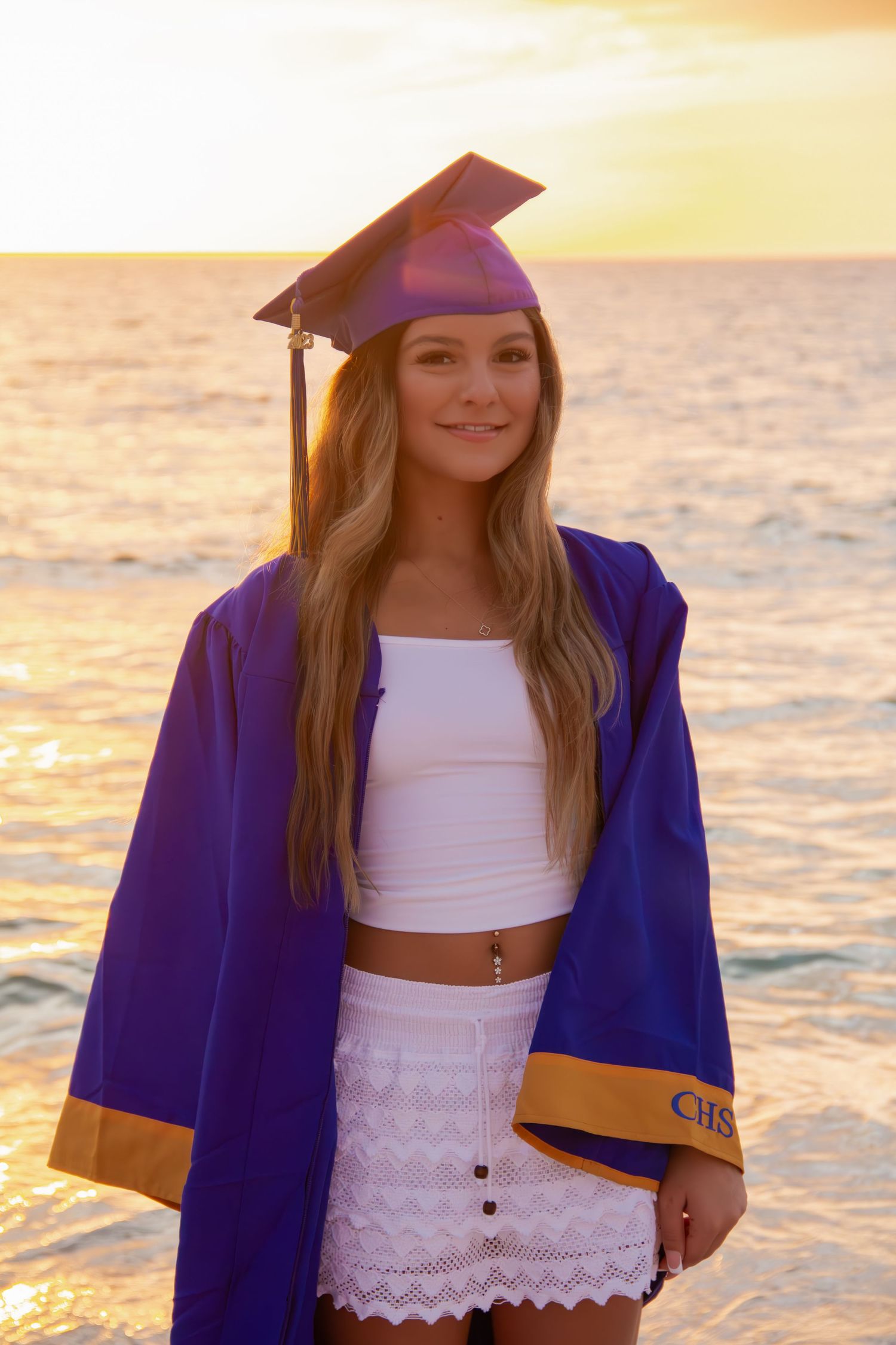 Young woman in purple graduation cap and gown posing on beach at golden sunset.