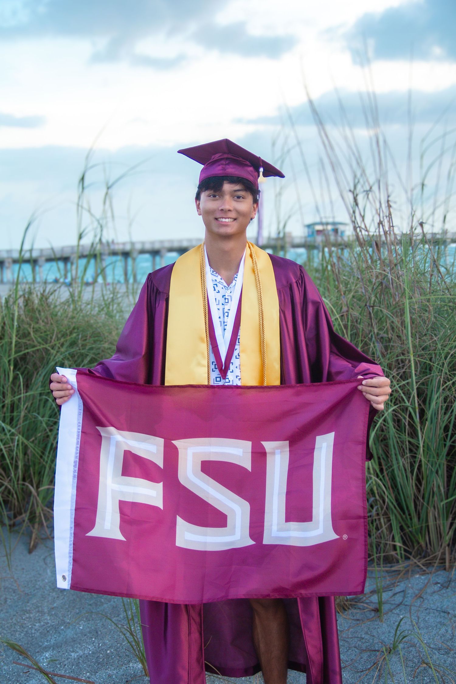 Graduate in maroon cap and gown holds FSU flag on beach, wearing gold honor stole and medal, smiling proudly.