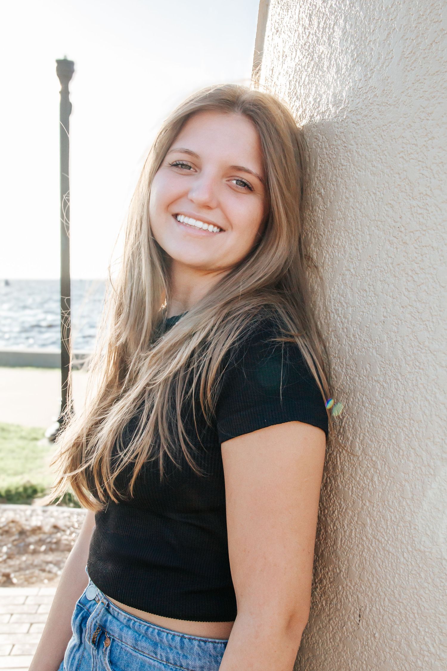 Smiling teenage girl with long hair leaning against a wall near the beach on a sunny day.